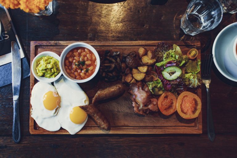Top view of delicious fried eggs and vegetables with sausages and beans on wooden tray with fork and knife